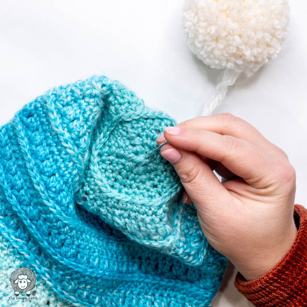 hands inserting a tapestry needle threaded onto the yarn strands of a pom pom through the center point of the top of a hat