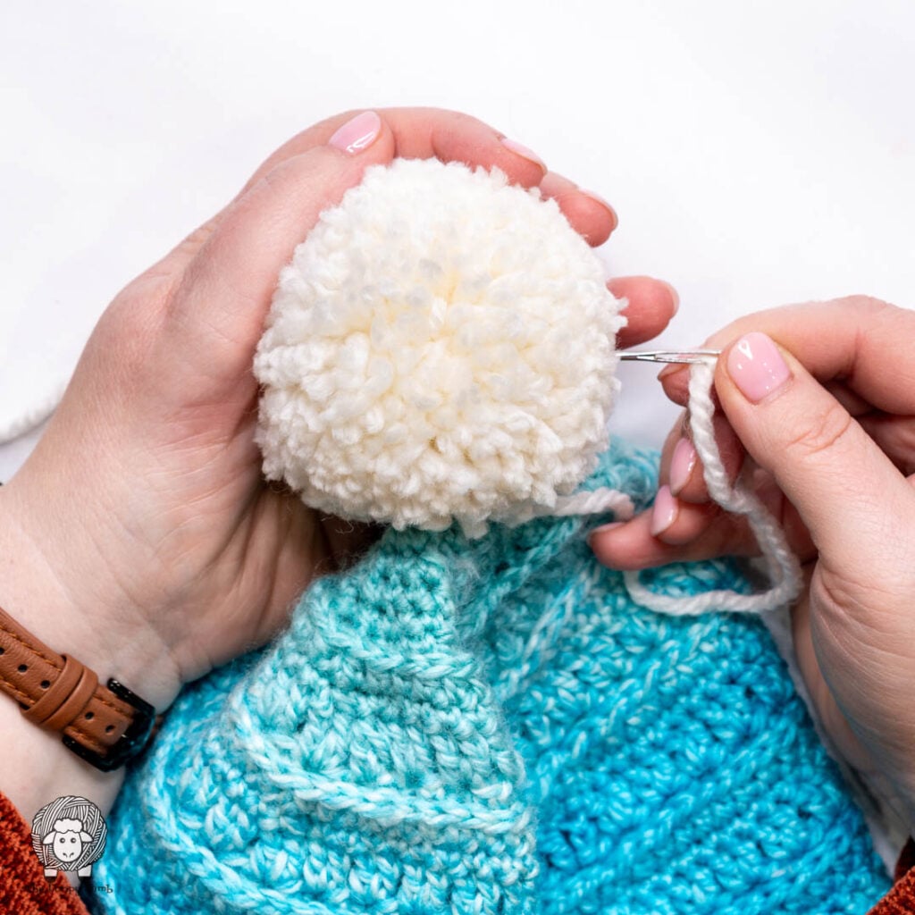 step three of how to attach a yarn pom pom to a hat - needle is being inserted through the center of the pom pom