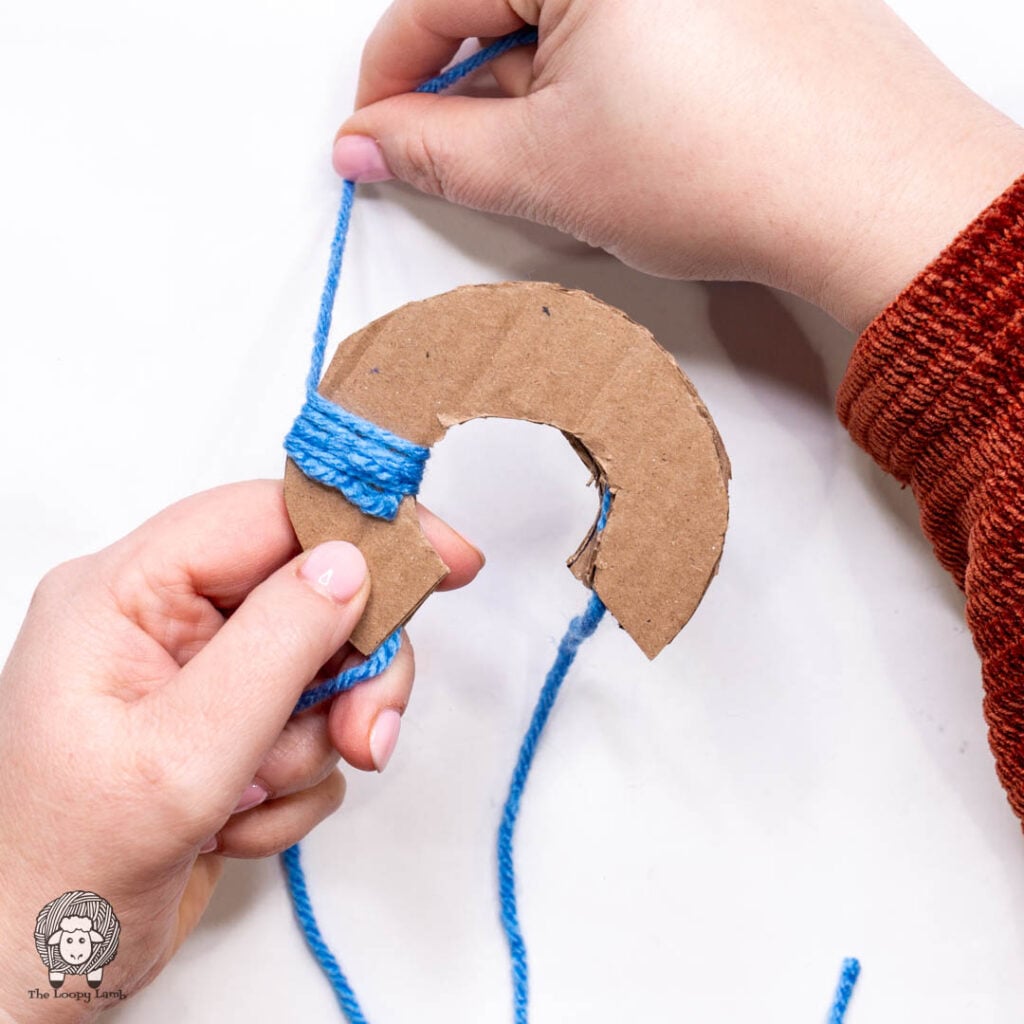 hands wrapping blue yarn around the cardboard pom pom maker