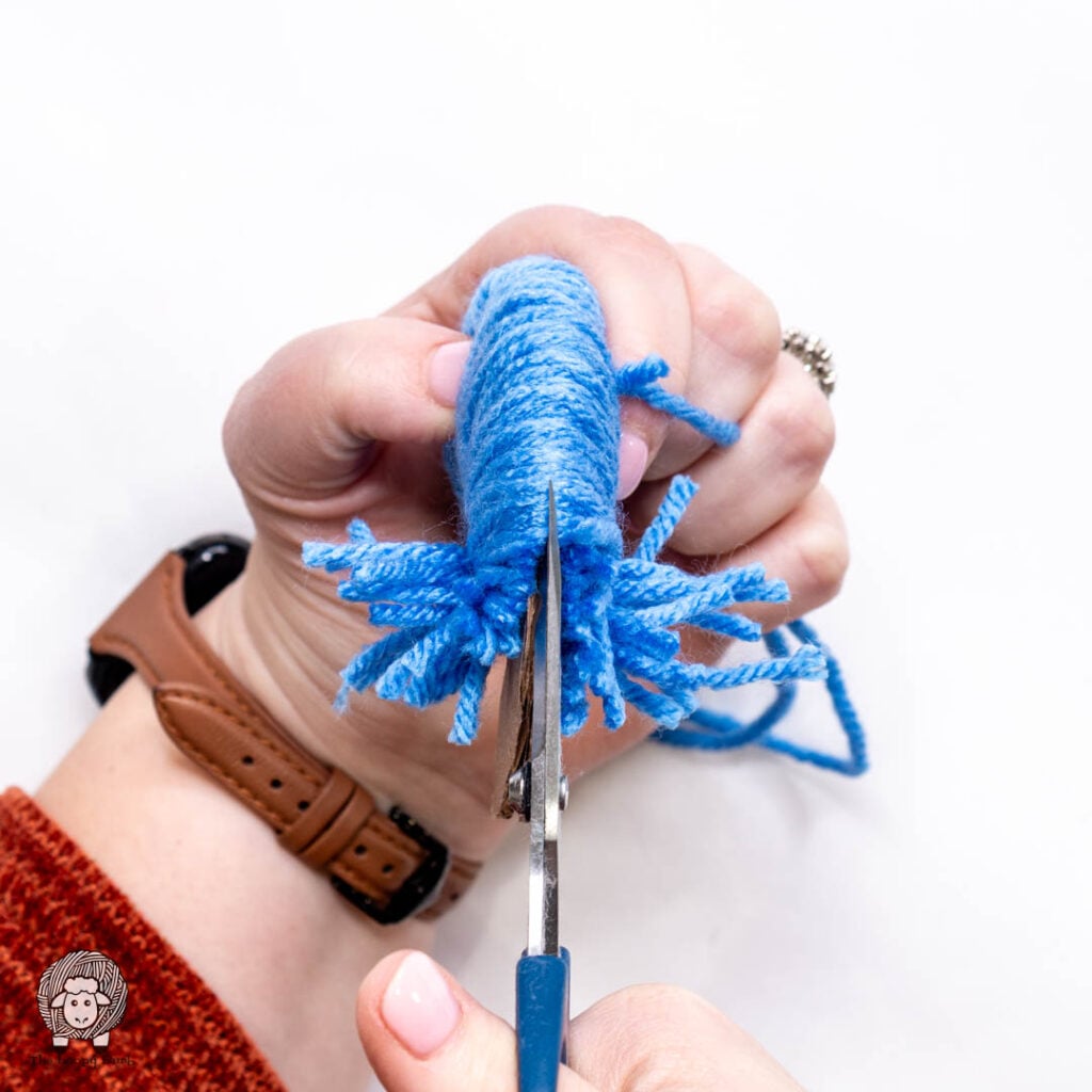 scissors being used to cut the wrapped yarn from the cardboard pom pom maker along the outside edge