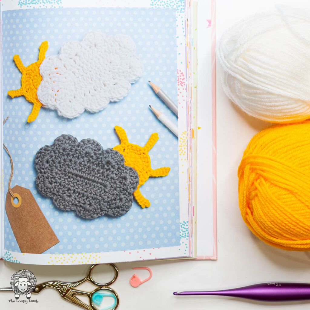 book open to show two different cloud and sun appliques next to a white and yellow balls of yarn