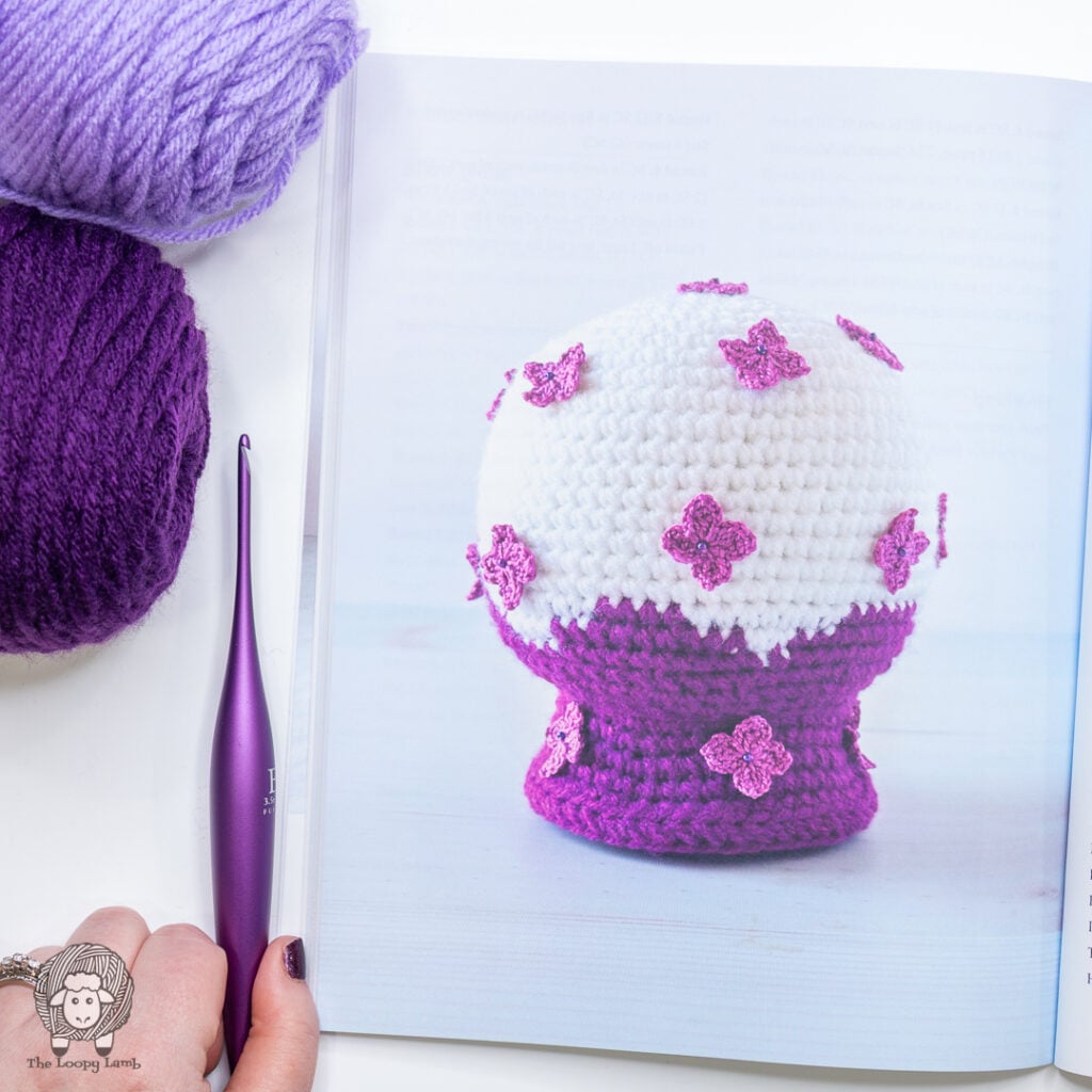 lilac themed crochet snow globe shown in the book next to a purple crochet hook and purple yarn