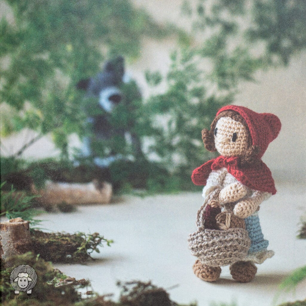 crochet red riding hook walking through the woods with the crochet woolf leering from a distance