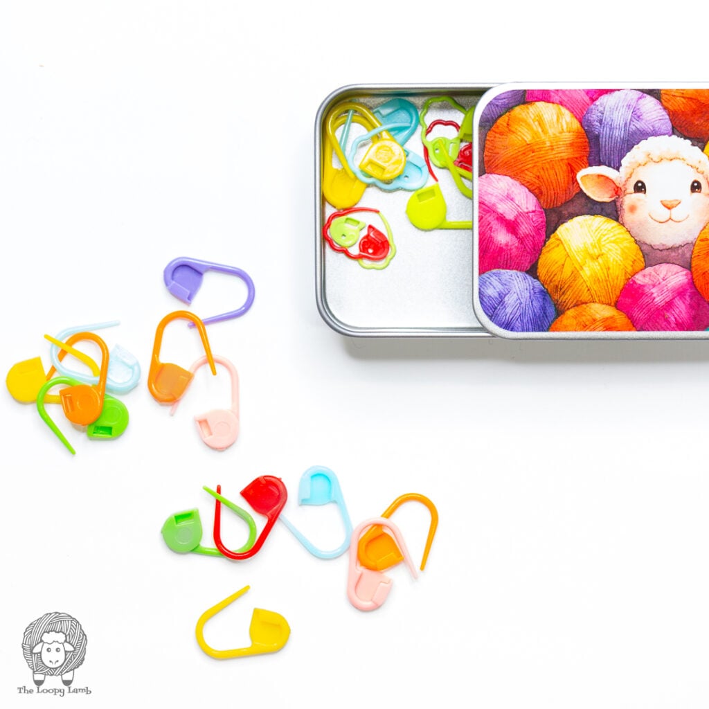 Colorful tin that's left open showing is houses colorful stitch markers that have spilled out onto a white surface
