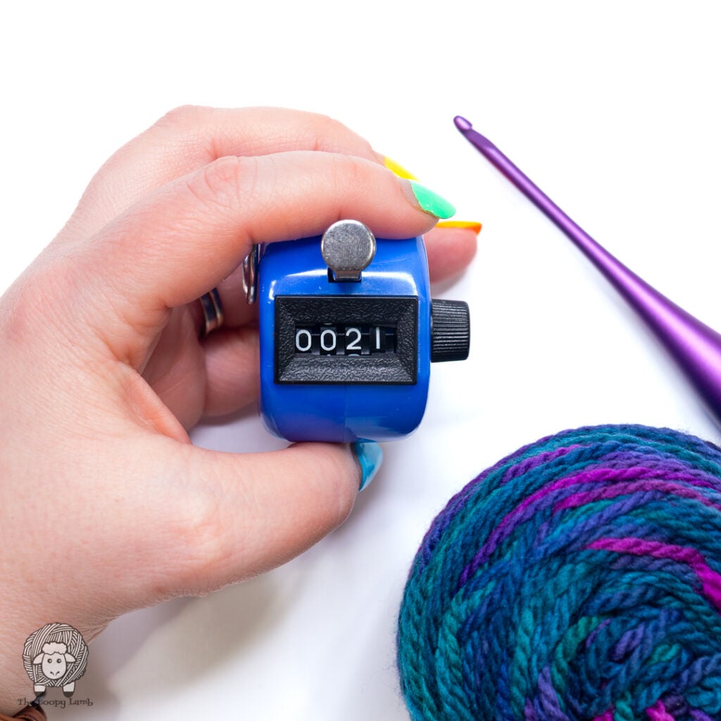 a manual row counter being held in a hand next to yarn and a purple crochet hook