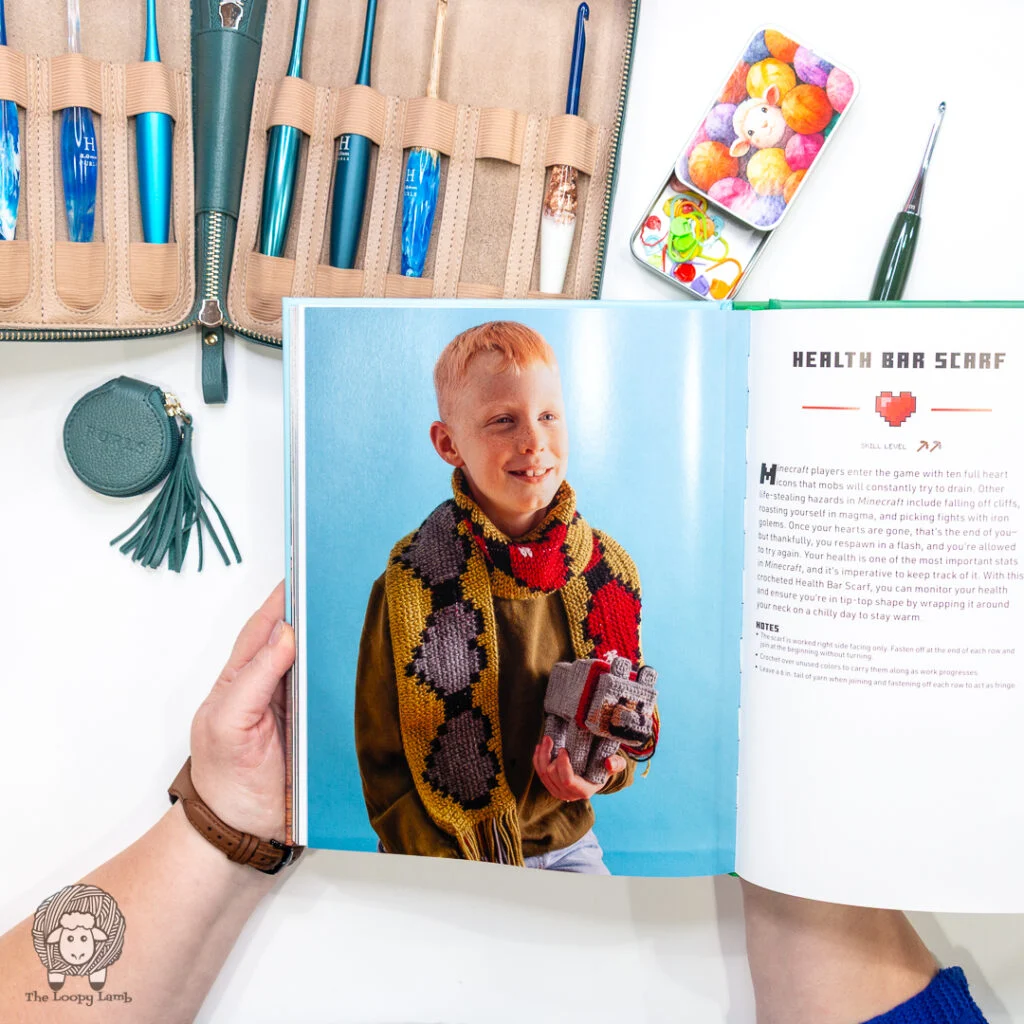 hands holding the Minecraft: the official crochet book open to show the Health Bar Scarf page surrounded by Furls crochet hooks & tools.