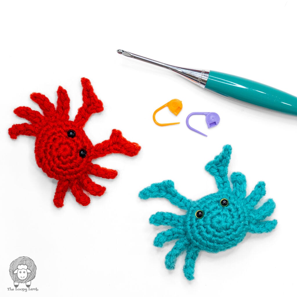 two mini no sew crochet crabs on a white backgroudn next to some stitch markers and a furls odyssey II crochet hook