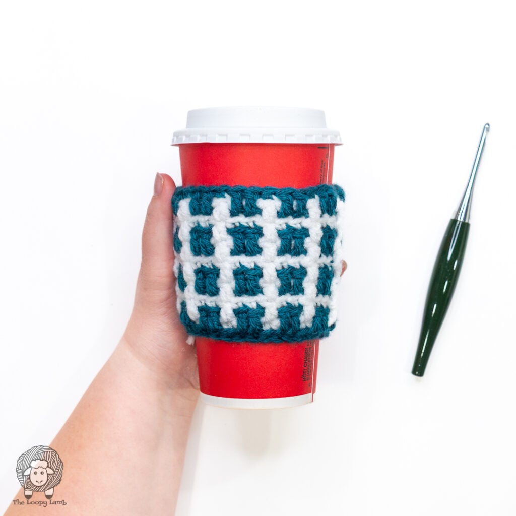 hand holding a teal and white crochet cup cozy against a white background next to a green crochet hook