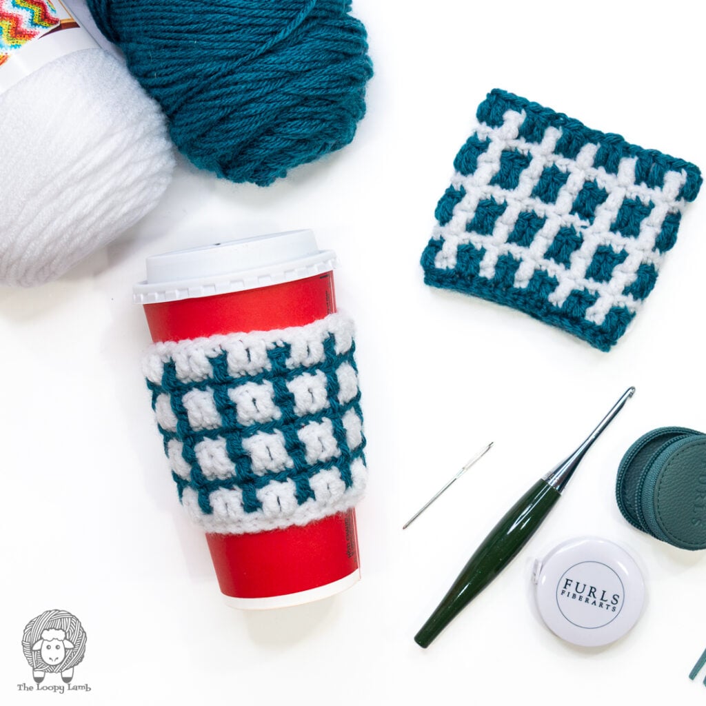 White and teal mosaic crochet cup cozy on a cup next to a teal and white cup cozy in a flat lay with yarn and a crochet hook